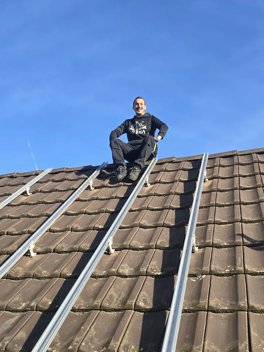 Braunes Ziegeldach von unten fotografiert: vier helle Montageschienen laufen bis zum First hinauf, oben auf dem First sitzt ein Monteur in schwarzer Arbeitskleidung