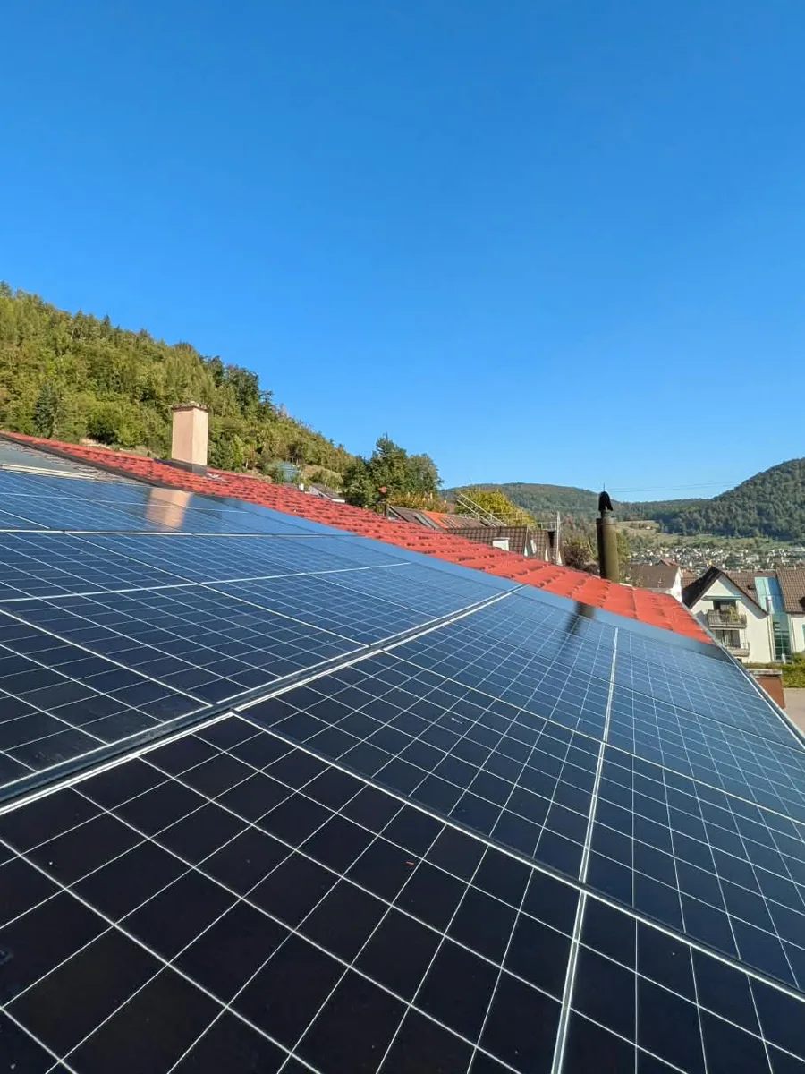 Schrägdach voller dunkler Solarmodule, von der Dachkante her fotografiert; daneben der rote Ziegelfirst, dahinter ein bewaldeter Hang und im Tal ein Dorf