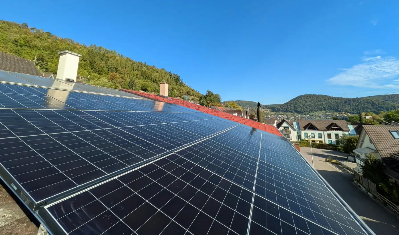 Schrägdach voller dunkler Solarmodule, von der Dachkante her fotografiert; daneben der rote Ziegelfirst, dahinter ein bewaldeter Hang und im Tal ein Dorf