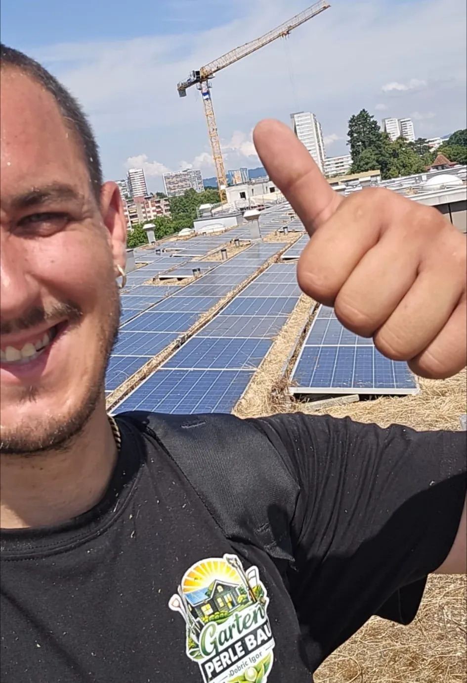 Igor Dobric auf einem Flachdach zwischen Reihen von Solarmodulen, ringsum trockenes Schnittgut, im schwarzen Firmen-T-Shirt mit dem Logo von Garten Perle Bau, lachend mit erhobenem Daumen