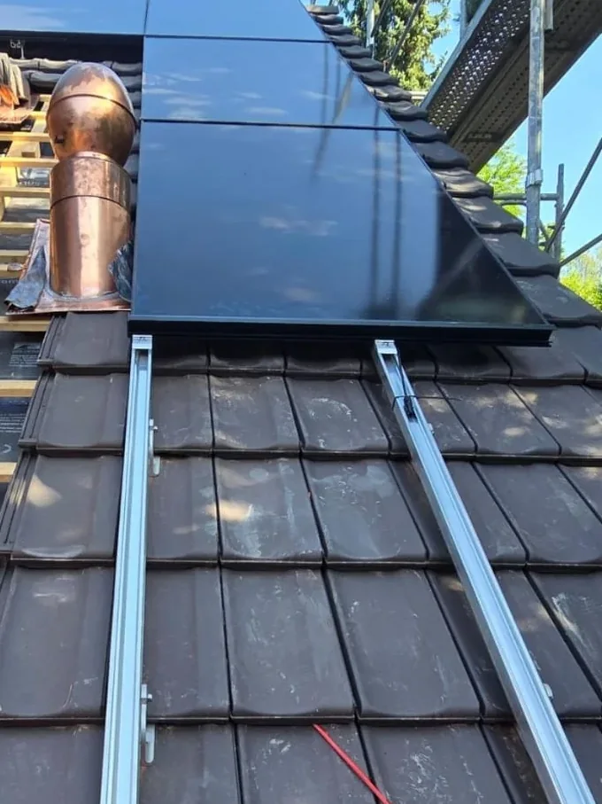 Solarmontage auf einem Ziegeldach: die Montageschienen sind gesetzt, das erste schwarze Modul liegt bereits auf, daneben das Gerüst und der Kupferkamin